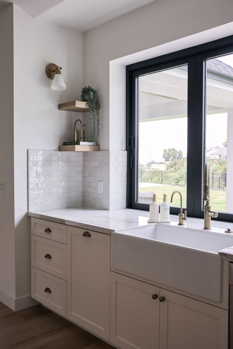 A bright kitchen featuring cream shaker-style cabinets with oil-rubbed bronze cup pulls, a white apron-front farmhouse sink, marble countertops, glossy white subway tile backsplash, floating wood shelves, and a black-framed window overlooking a backyard.