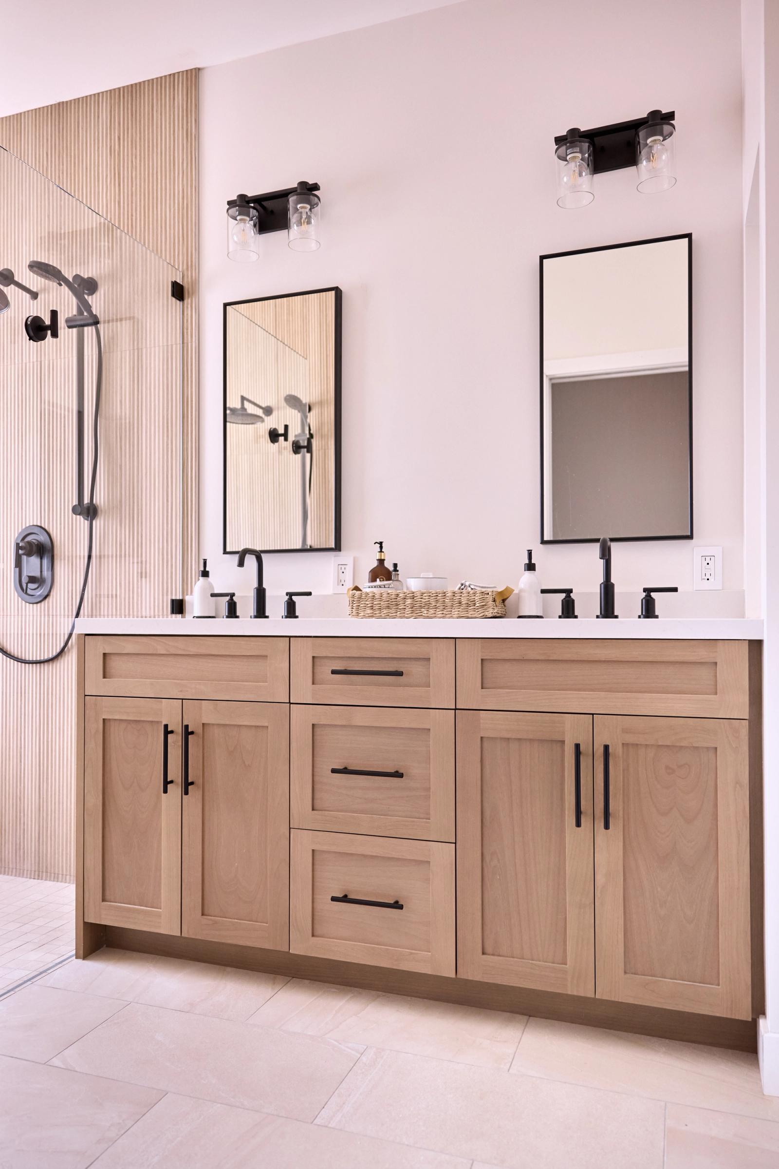 A modern double bathroom vanity with light natural wood shaker-style cabinets, white countertop, matte black faucets and hardware, two rectangular black-framed mirrors, and dual wall sconces. A walk-in shower with fluted tile is visible to the left.