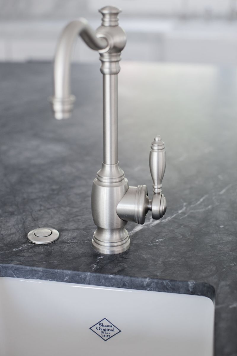 A close-up of a traditional-style brushed nickel faucet and lever handle mounted on a dark soapstone countertop above a Shaw's Original farmhouse sink.