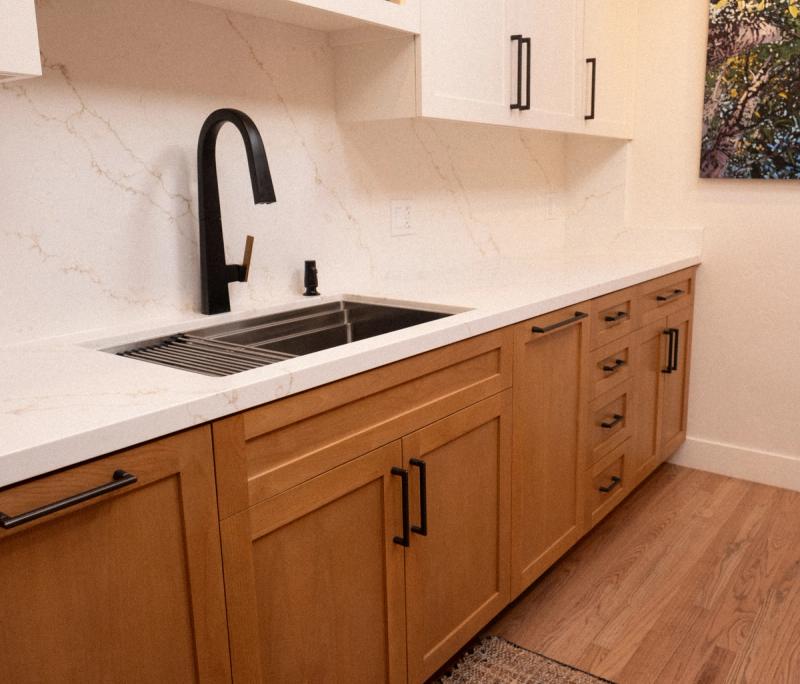 A modern kitchen featuring warm white oak shaker-style lower cabinets and white upper cabinets, topped with white quartz countertops and backsplash. A matte black pull-down faucet is mounted above an undermount sink with a roll-up drying rack.