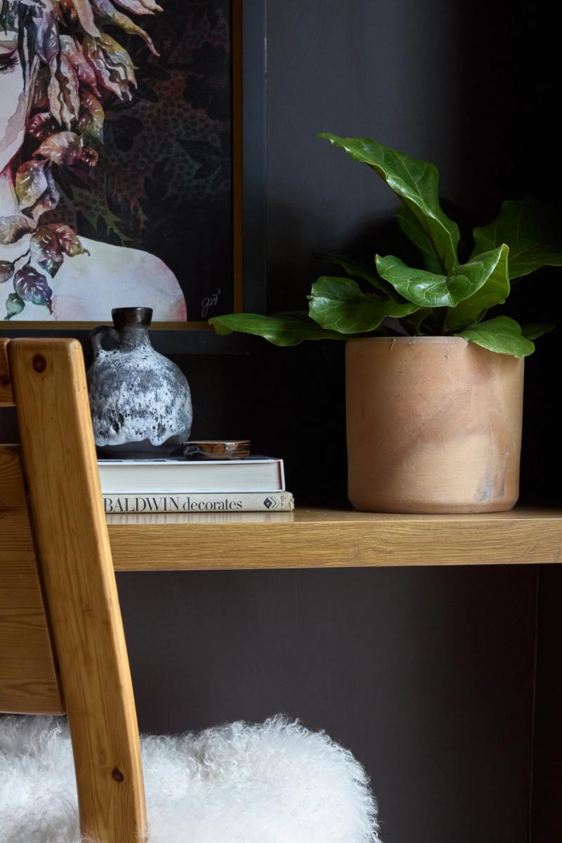 A close-up of a warm oak wood shelf styled with a terracotta pot holding a fiddle leaf fig, a ceramic vase, and a stack of books including 'Baldwin Decorates,' set against a dark charcoal wall with framed artwork in the background.