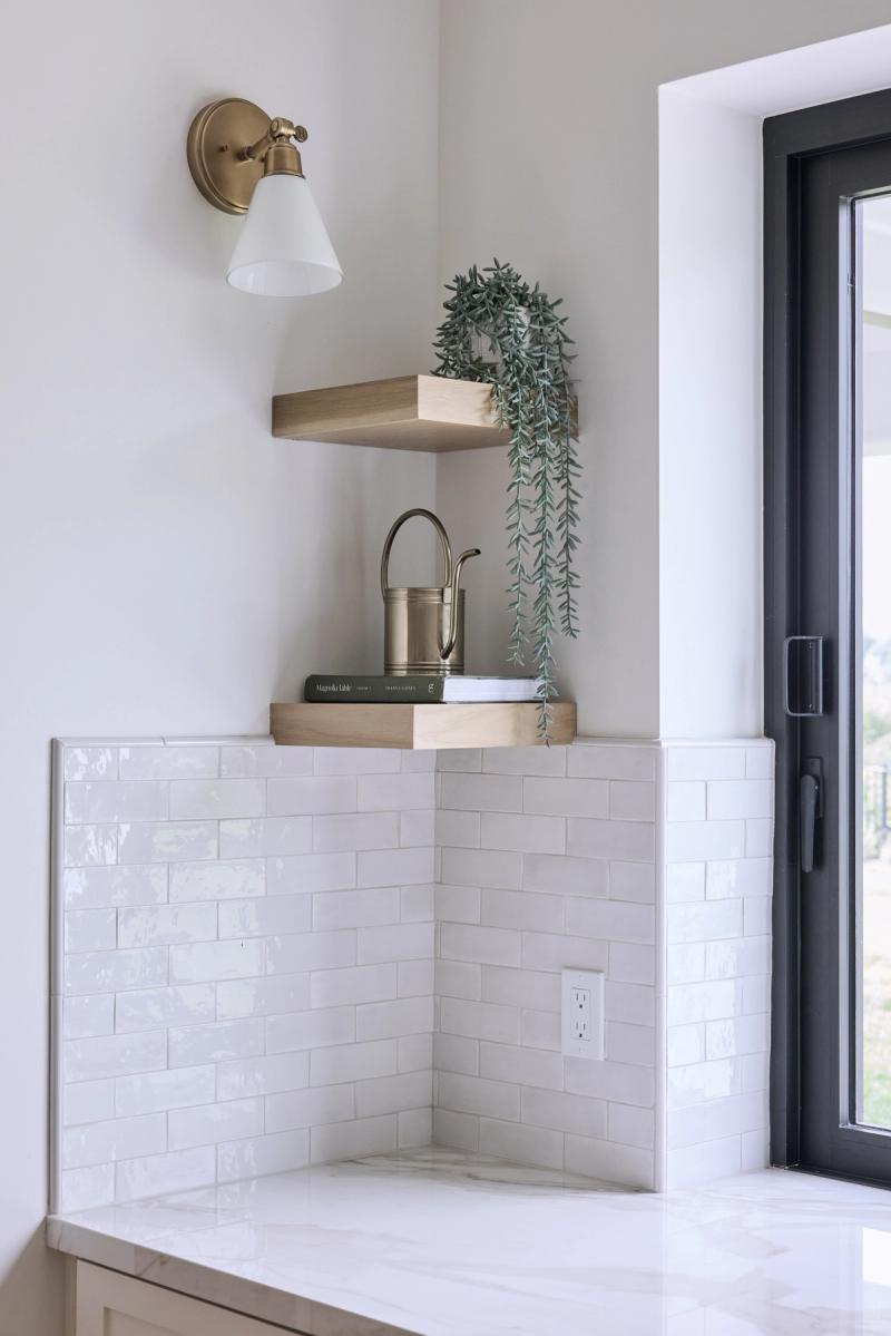 A bright kitchen corner featuring glossy white subway tile backsplash, light wood floating shelves styled with books and a trailing plant, a brass wall sconce, and a marble countertop beside a black-framed glass door.