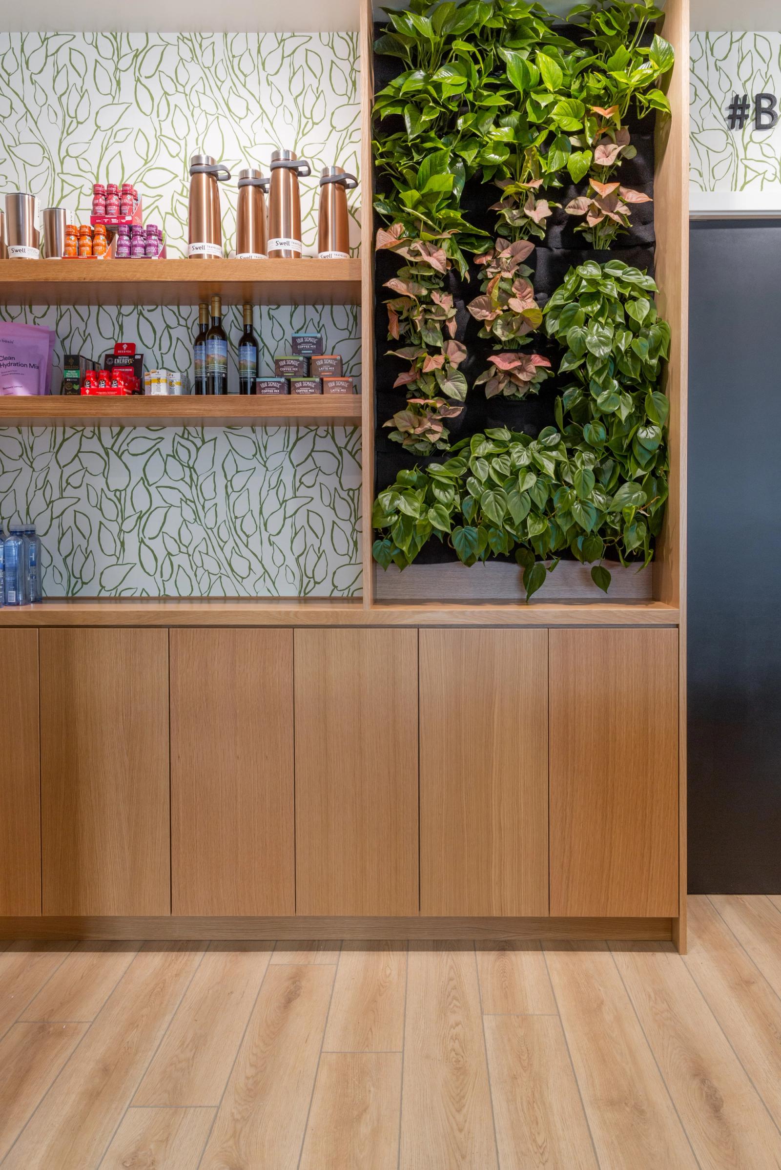 A modern built-in wooden shelving unit with copper water bottles, beverages, and packaged goods displayed against a leaf-patterned wallpaper, beside a vertical living plant wall featuring pothos and syngonium plants, all set into warm oak cabinetry.
