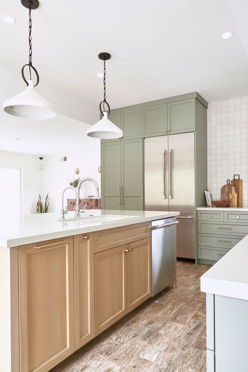 A bright, modern-farmhouse kitchen featuring sage green shaker-style perimeter cabinets, a natural white oak island with white quartz countertops, stainless steel appliances, and white pendant lights hanging from a white ceiling.