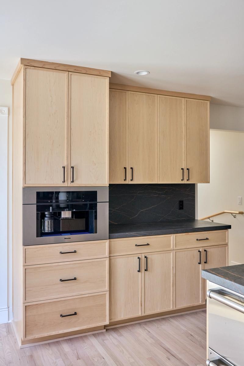 A modern kitchen wall unit featuring light maple cabinetry with black bar pulls, a built-in Miele coffee machine, dark soapstone countertop and backsplash, and multiple upper and lower cabinets with drawers.