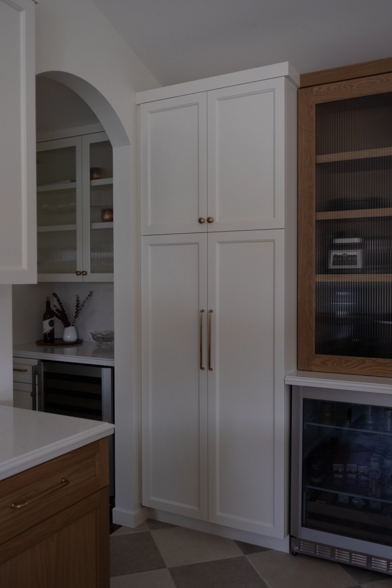A transitional kitchen featuring a tall white shaker-style pantry cabinet with brass hardware, warm wood lower cabinets, a wine refrigerator, and an arched butler's pantry alcove with ribbed glass cabinet doors in the background.