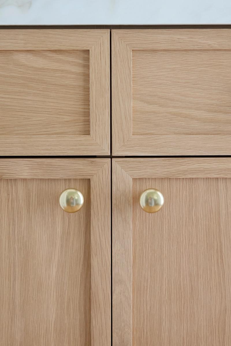 Close-up of light natural oak shaker-style cabinet doors with recessed panels and round polished brass knobs, topped with a white marble countertop.