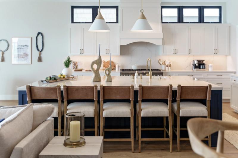 A bright, open-concept kitchen featuring white shaker cabinets with brass hardware, a navy blue island with white quartz countertop, leather and linen bar stools, and two white cone pendant lights. The space flows into a living area and includes a marble slab backsplash and professional-grade range.