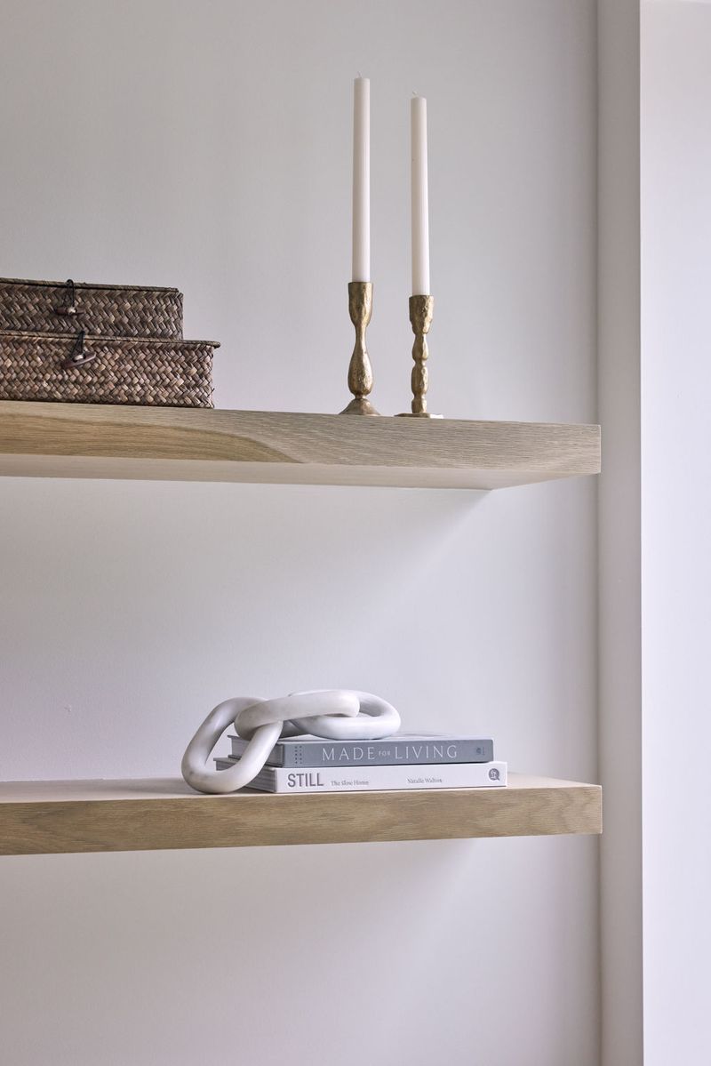 Two floating white oak shelves mounted on a white wall, styled with brass candlesticks, a woven basket, stacked coffee table books, and a white chain sculpture.