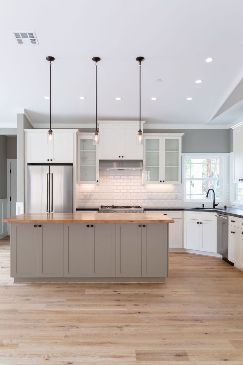 A bright, open kitchen featuring white shaker upper cabinets, a gray-green island with a butcher block countertop, white subway tile backsplash, stainless steel appliances, and light hardwood floors. Three glass pendant lights hang above the large center island.