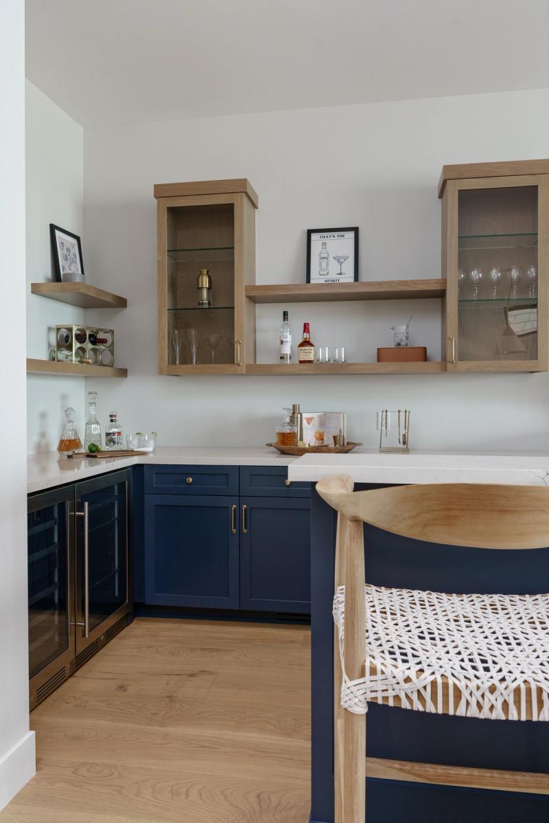 A stylish home bar featuring navy blue shaker-style lower cabinets with brass hardware, white quartz countertops, natural white oak upper cabinets and floating shelves, stocked with glassware and spirits, with a wine refrigerator on the left.