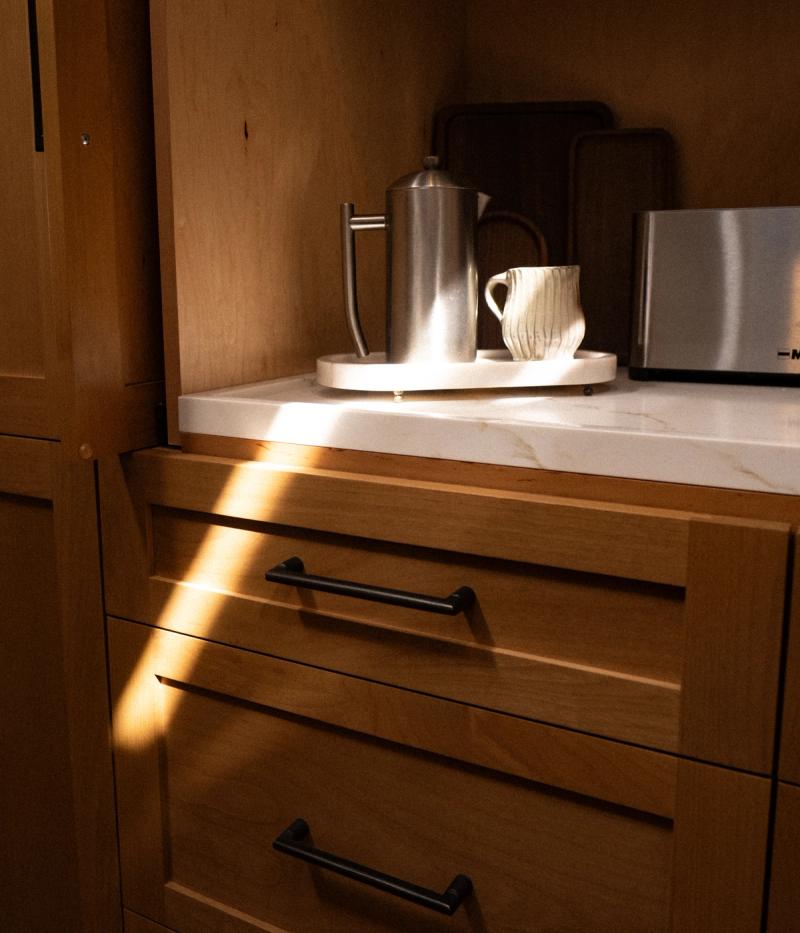 Close-up of custom natural wood kitchen drawers with matte black bar pulls and a white marble countertop, featuring a stainless steel French press and ceramic mug on a round tray, lit by warm natural light.