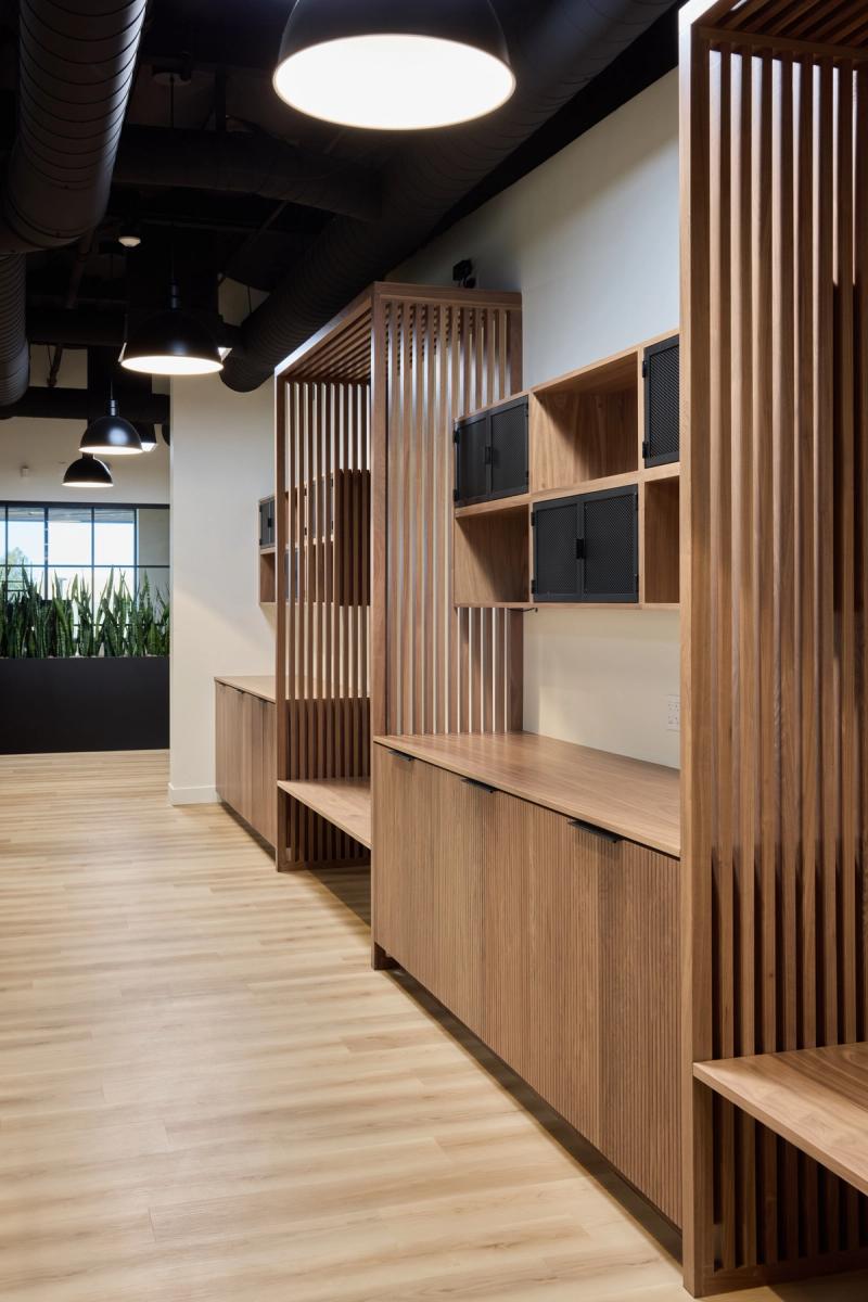 A row of warm-toned wood built-in cabinets and shelving units with vertical slatted dividers and black mesh cabinet doors, installed along a hallway in a modern office space with light hardwood floors and an exposed black ceiling.