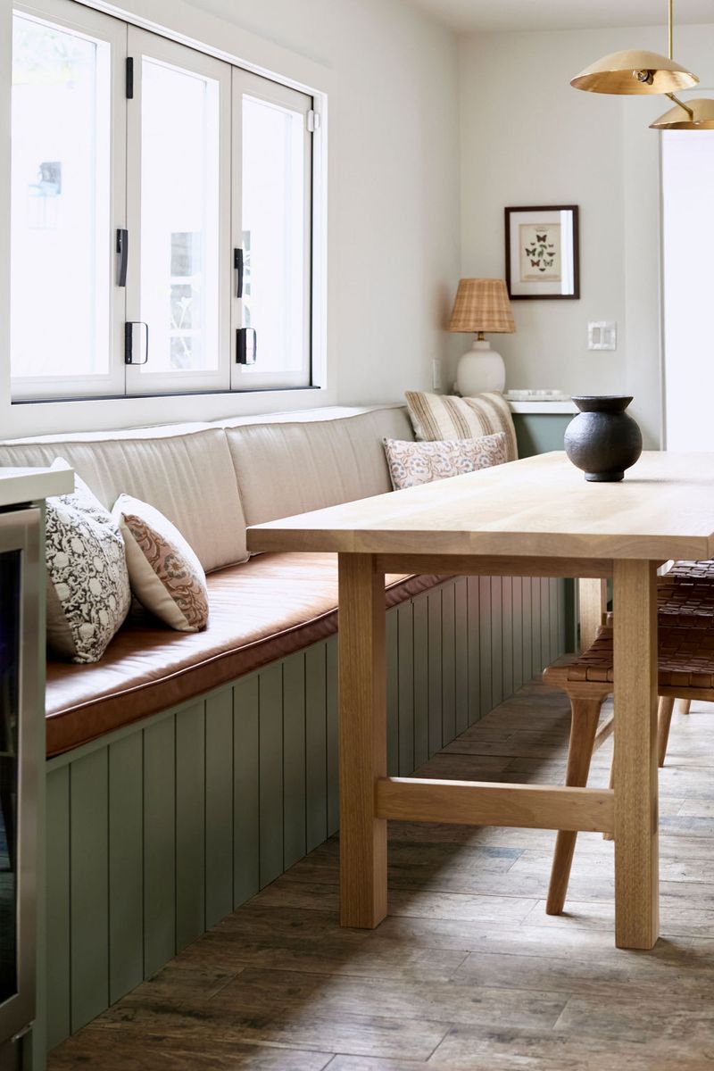 A built-in dining banquette with sage green beadboard paneling and a cognac leather cushion wraps around a corner, paired with a light oak dining table and decorative throw pillows near a bright window.
