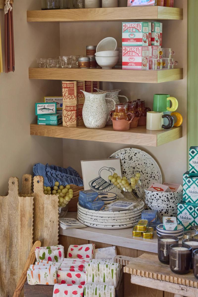 Floating light oak shelves mounted in a corner display an assortment of artisan food products, ceramics, mugs, and glassware above a counter styled with speckled plates, bowls, wooden cutting boards, and decorative kitchen goods.