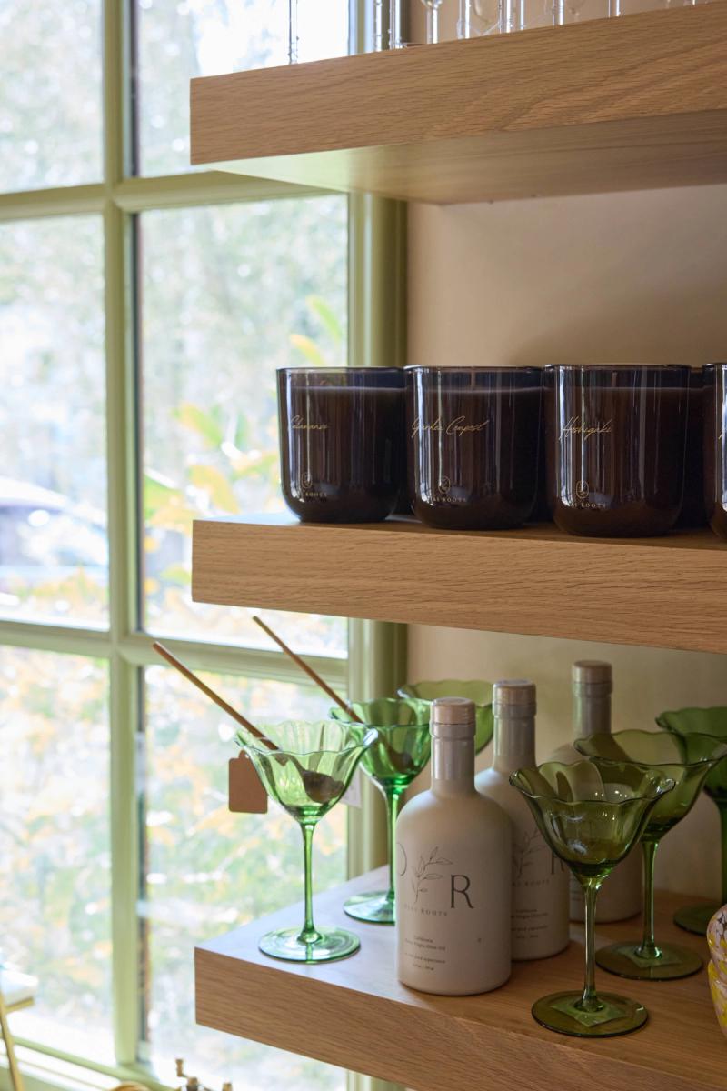 Close-up of light oak floating shelves mounted against a window, styled with dark amber candles labeled 'Roots,' green martini glasses, and white ceramic bottles with botanical branding.