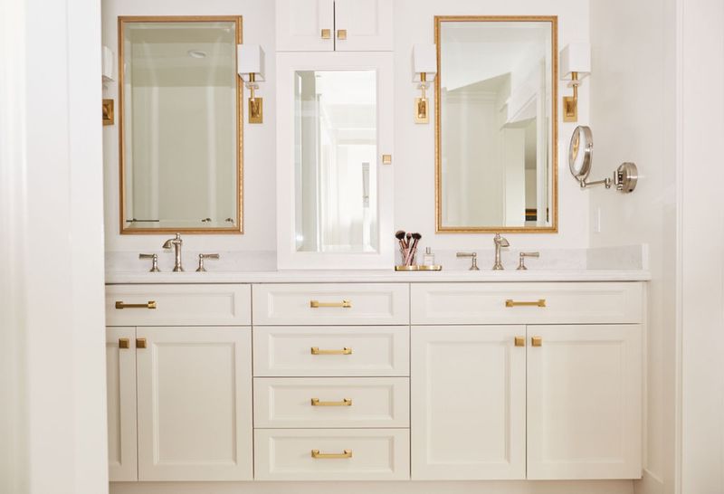 A bright, white double bathroom vanity with raised-panel cabinet doors, gold hardware, two gold-framed mirrors, and wall sconces flanking a center medicine cabinet.