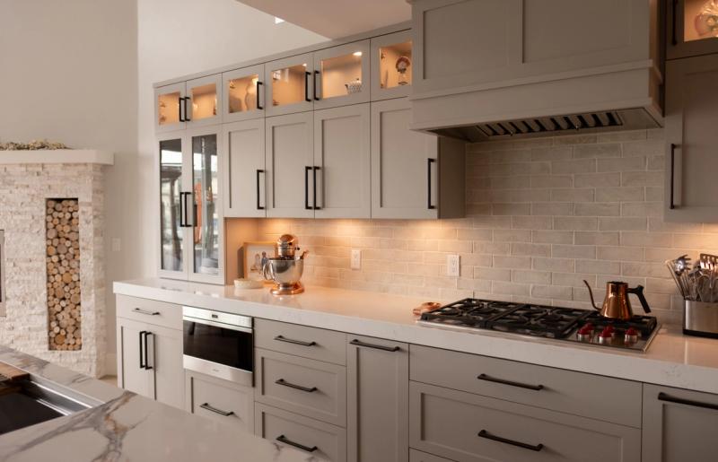 A custom kitchen featuring greige shaker-style cabinets with matte black hardware, a natural stone brick backsplash, stainless gas cooktop with range hood, and warm under-cabinet lighting. A decorative wood and stone fireplace surround is visible on the left.