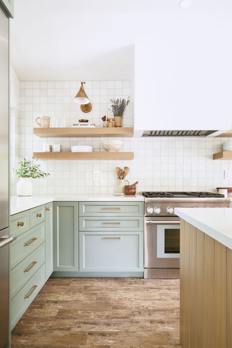 A bright, airy kitchen featuring sage green shaker-style lower cabinets with brass hardware, white square tile backsplash, open wood floating shelves, and a stainless steel range. The space is styled with natural wood accents and greenery.