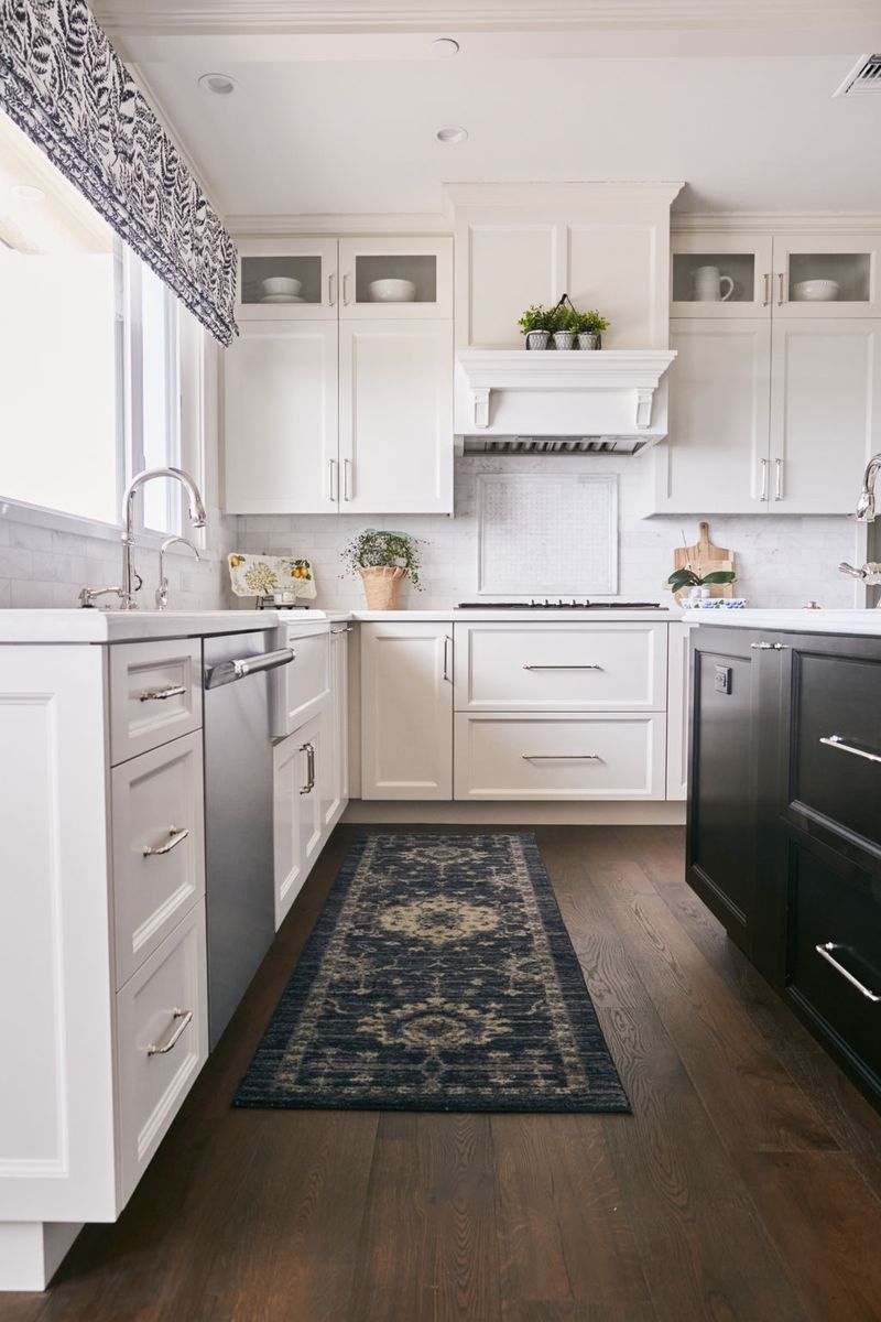 A bright, traditional-style kitchen featuring white inset cabinetry with nickel hardware, a custom range hood with corbels, dark hardwood floors, a navy runner rug, and a farmhouse sink beneath a window with a blue and white Roman shade.