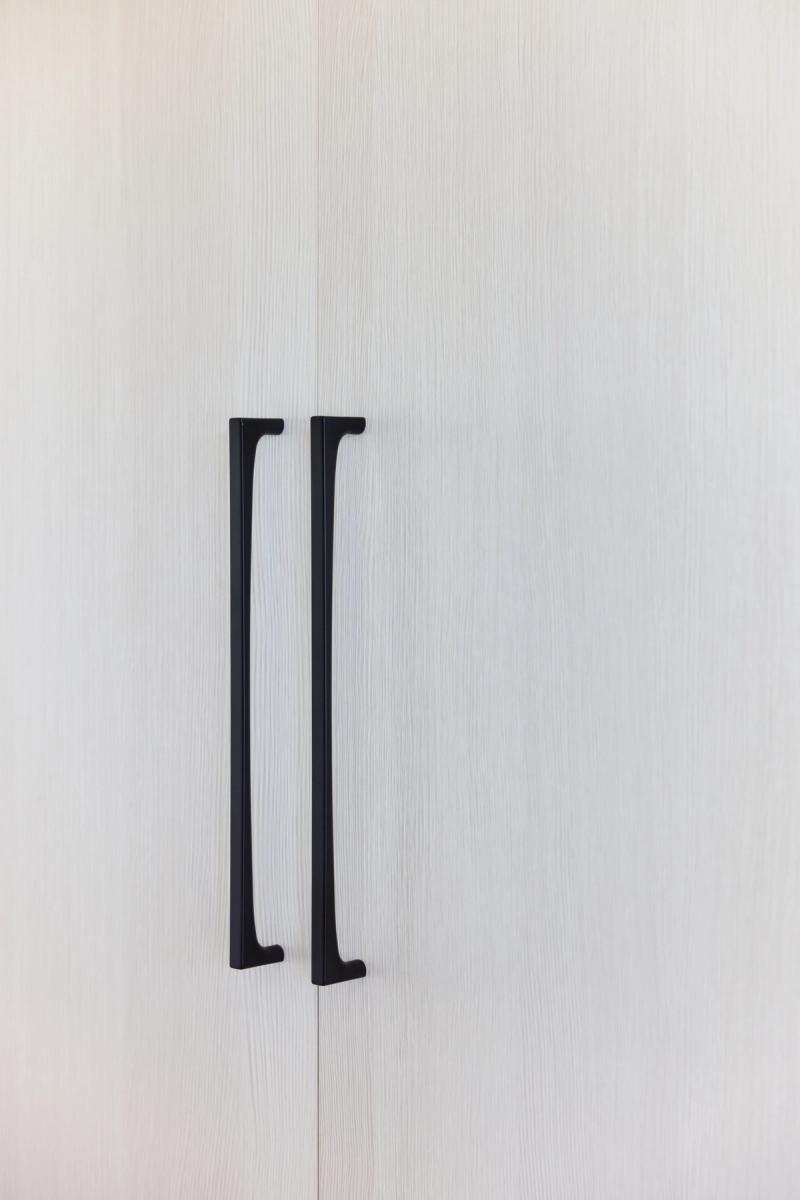 A close-up of two sleek matte black bar pulls mounted on light white oak cabinet doors, shown at the seam where two doors meet.