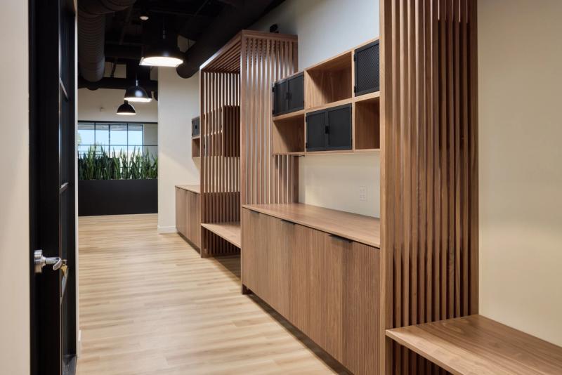 A custom walnut built-in cabinetry unit featuring vertical slatted wood dividers, open shelving with mesh-door cabinets, and lower fluted-front cabinets, installed along a hallway with light hardwood floors and an exposed black ceiling beyond.