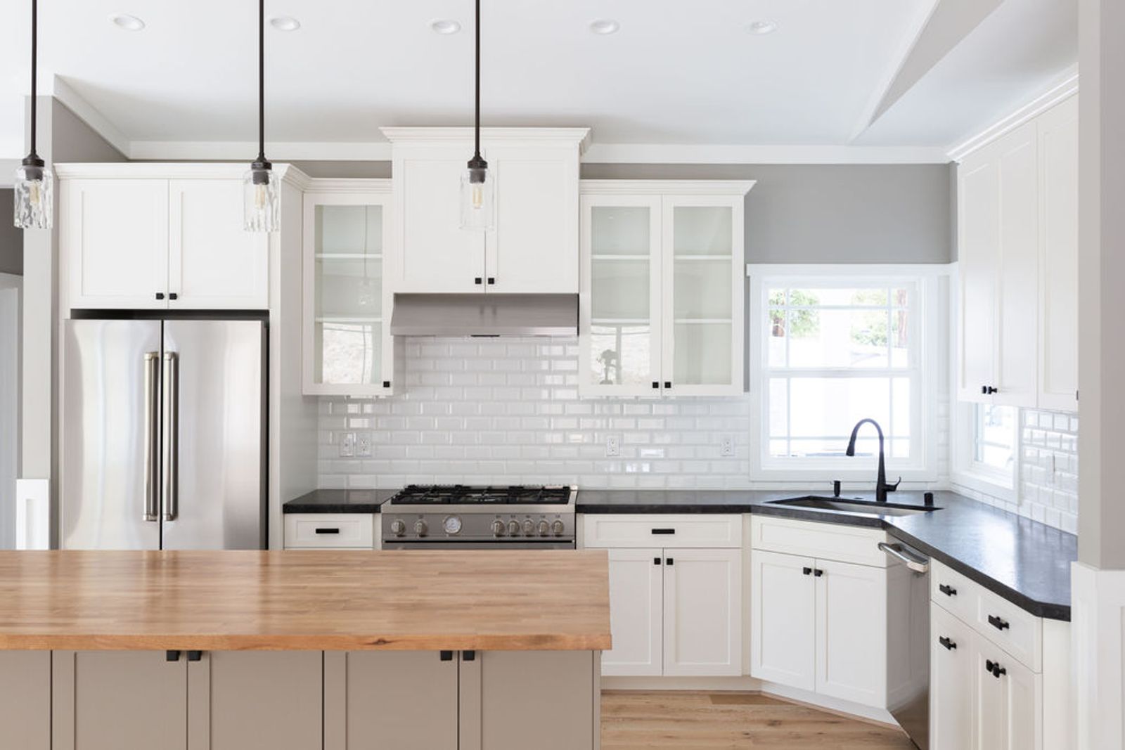 A bright, newly completed kitchen featuring white shaker cabinets, black granite countertops, a white subway tile backsplash, stainless steel appliances, and a butcher block island with a taupe base.