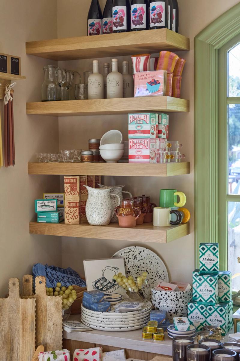 A set of light oak floating corner shelves styled with gourmet food products, glassware, ceramics, tea boxes, and wine bottles in a bright boutique shop setting.