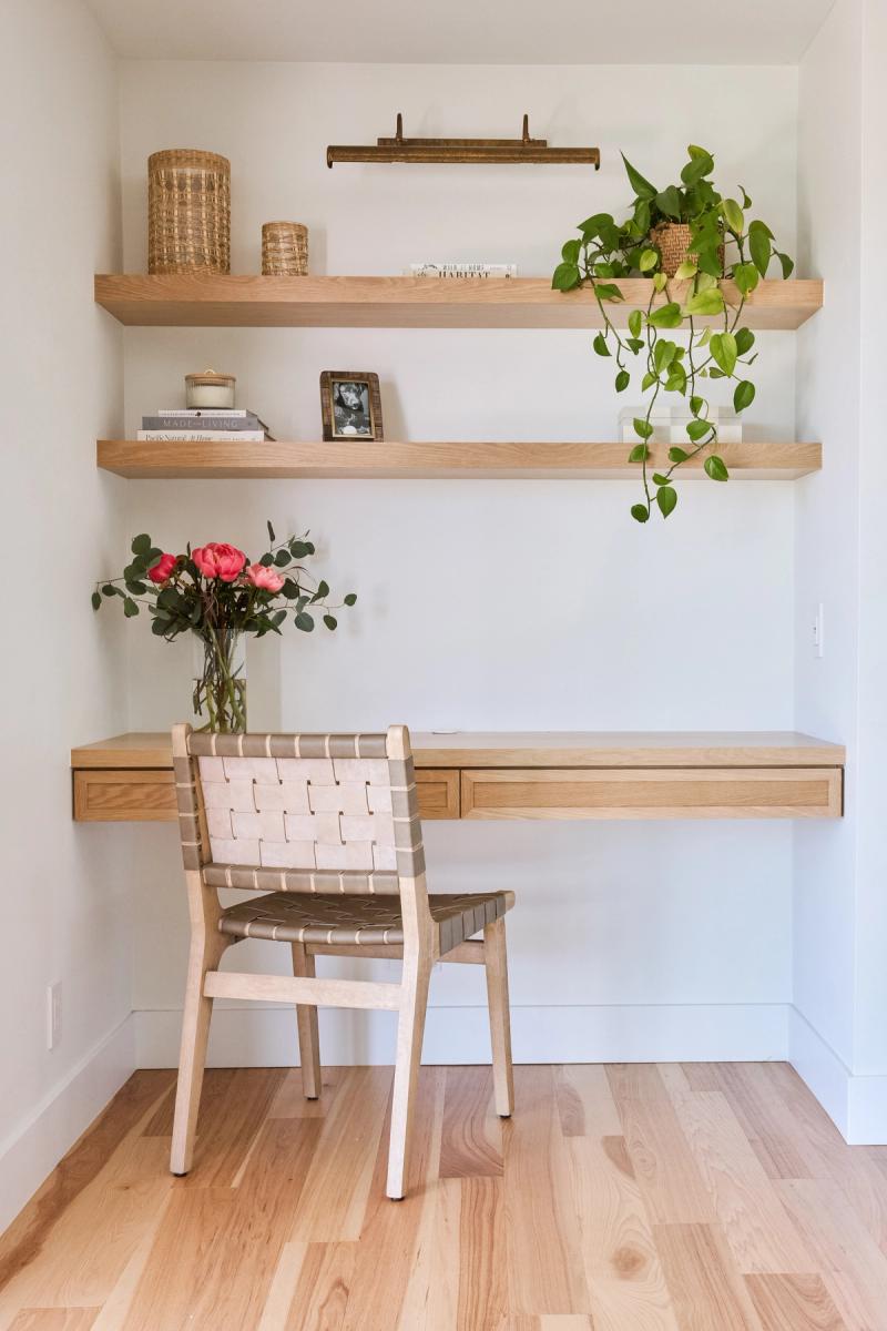 A built-in home office nook featuring floating white oak shelves and a wall-mounted desk with drawer, styled with books, woven baskets, a trailing pothos plant, and a vase of pink roses, paired with a woven leather chair.