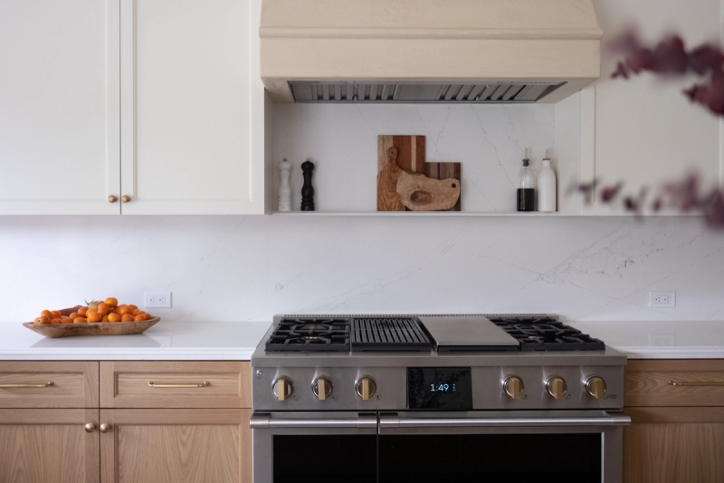 A bright, modern transitional kitchen featuring white upper cabinets, natural oak lower cabinets with brass hardware, a stainless steel professional range with gold knobs, and a white marble-look quartz backsplash and countertops.