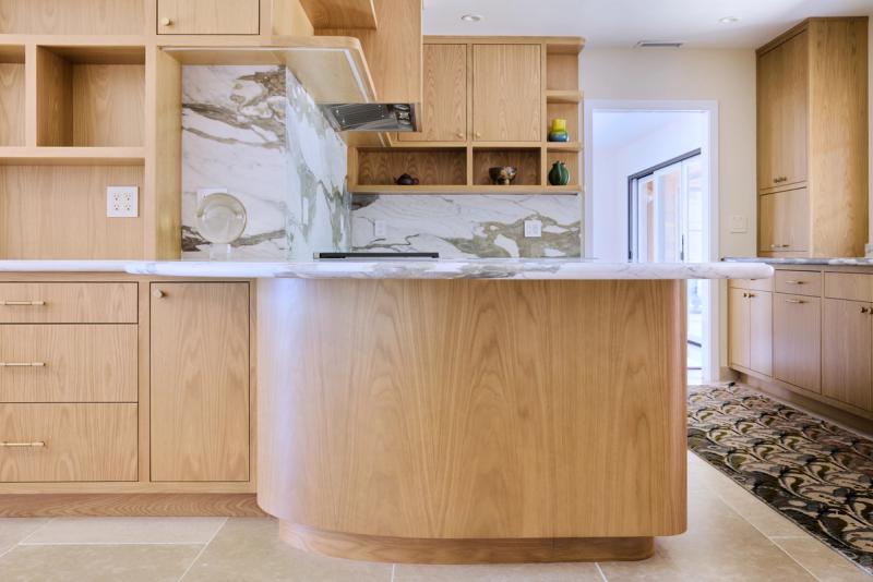 A custom kitchen featuring light natural oak cabinetry with a curved island topped with white and green marble, open shelving, and a dramatic full-height marble backsplash behind the cooktop.