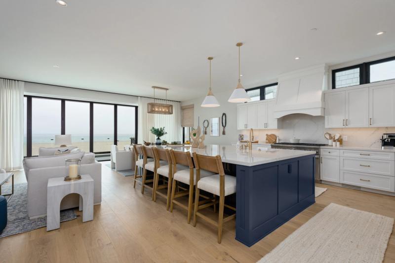 A bright open-concept coastal kitchen featuring a large navy blue island with white quartz countertop and bar seating, white shaker cabinets with gold hardware, and floor-to-ceiling windows overlooking the ocean.