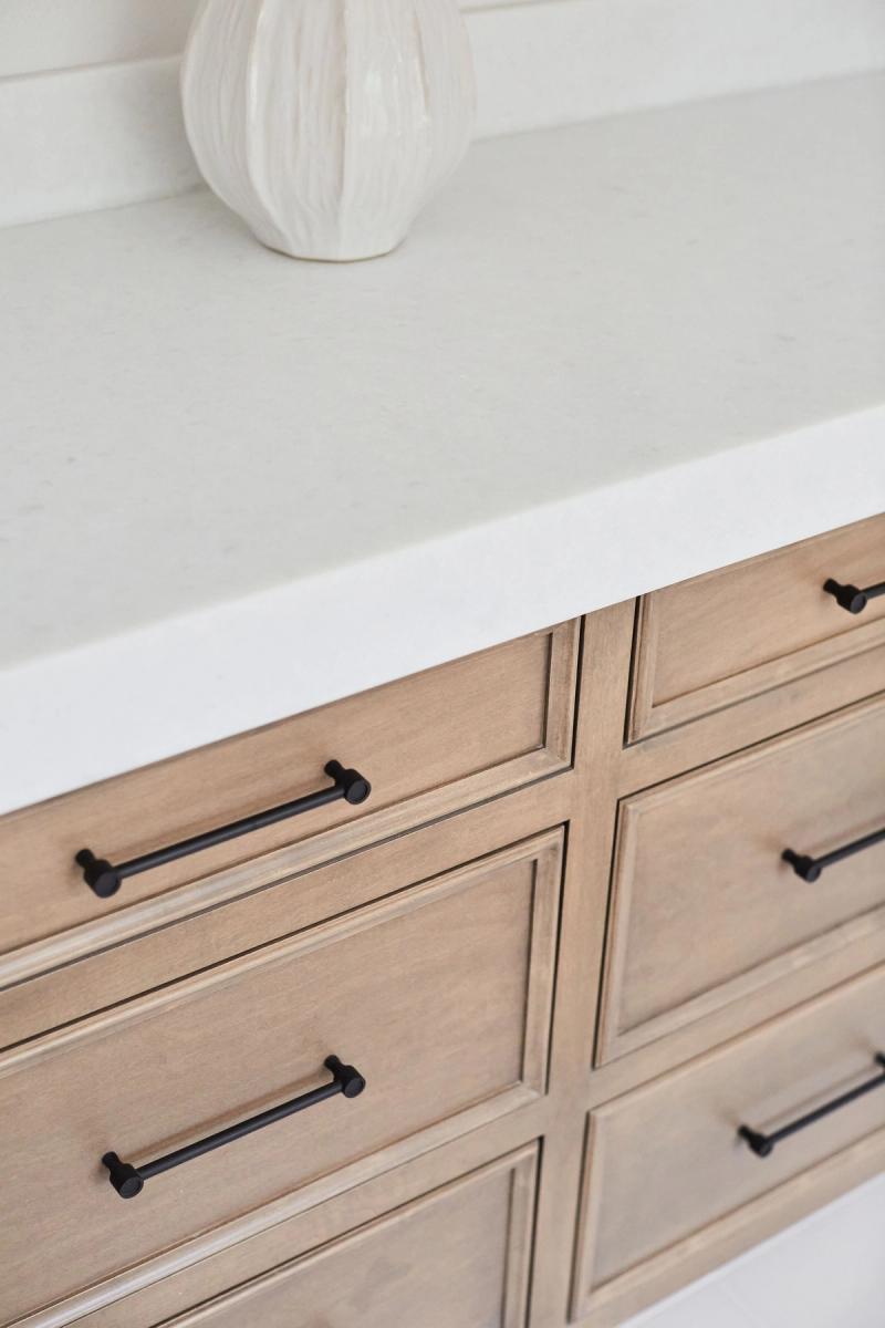 A close-up of a light wood bathroom vanity with raised panel drawer fronts, matte black bar pulls, a white marble countertop, and a decorative white ceramic vase in the background.