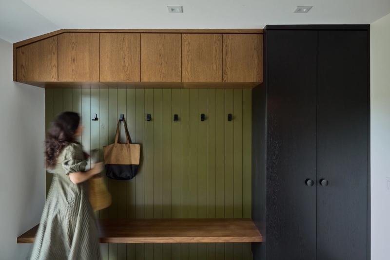 A custom mudroom built-in featuring warm oak upper cabinets and bench, an olive green vertical shiplap wall with black coat hooks, and a dark oak wardrobe cabinet to the right. A woman in motion reaches for a hanging tote bag.