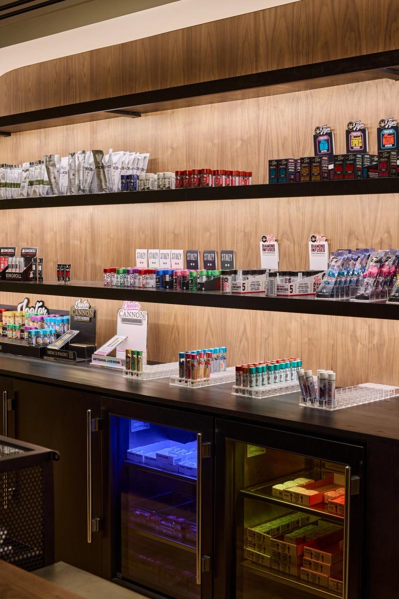 A modern retail display wall featuring warm wood shelving with built-in lighting, displaying packaged products on multiple levels, with refrigerated display cases below the counter.