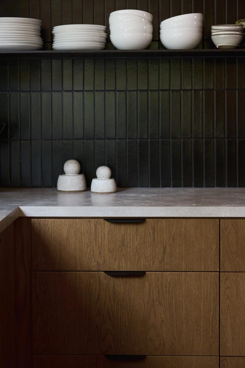 A modern kitchen featuring warm oak wood drawers with dark metal pulls, a light stone countertop, and a deep green vertical tile backsplash with open shelving holding stacked white dishes and bowls.