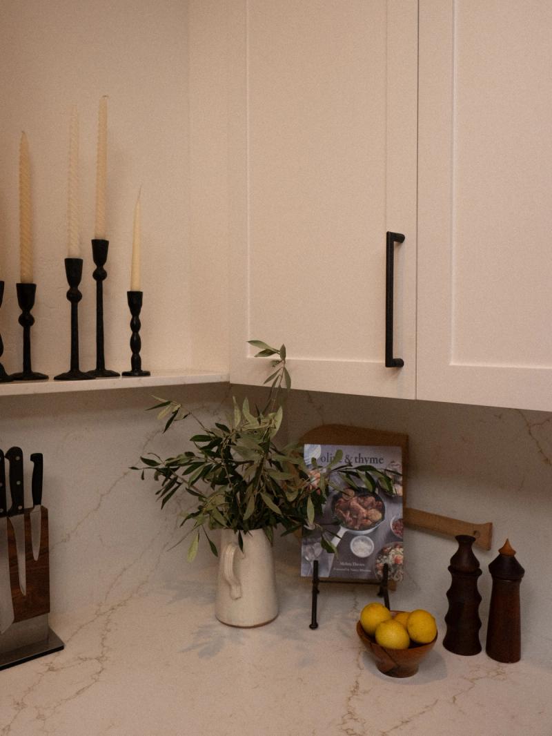 A styled kitchen corner featuring white shaker cabinets with matte black bar pulls, a marble countertop, black candlestick holders, an olive branch arrangement in a ceramic pitcher, and a wooden bowl of lemons.