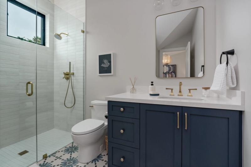 A modern bathroom featuring a navy blue shaker-style vanity with brass hardware and white quartz countertop, paired with a glass-enclosed shower with white subway tile and a patterned mosaic floor.
