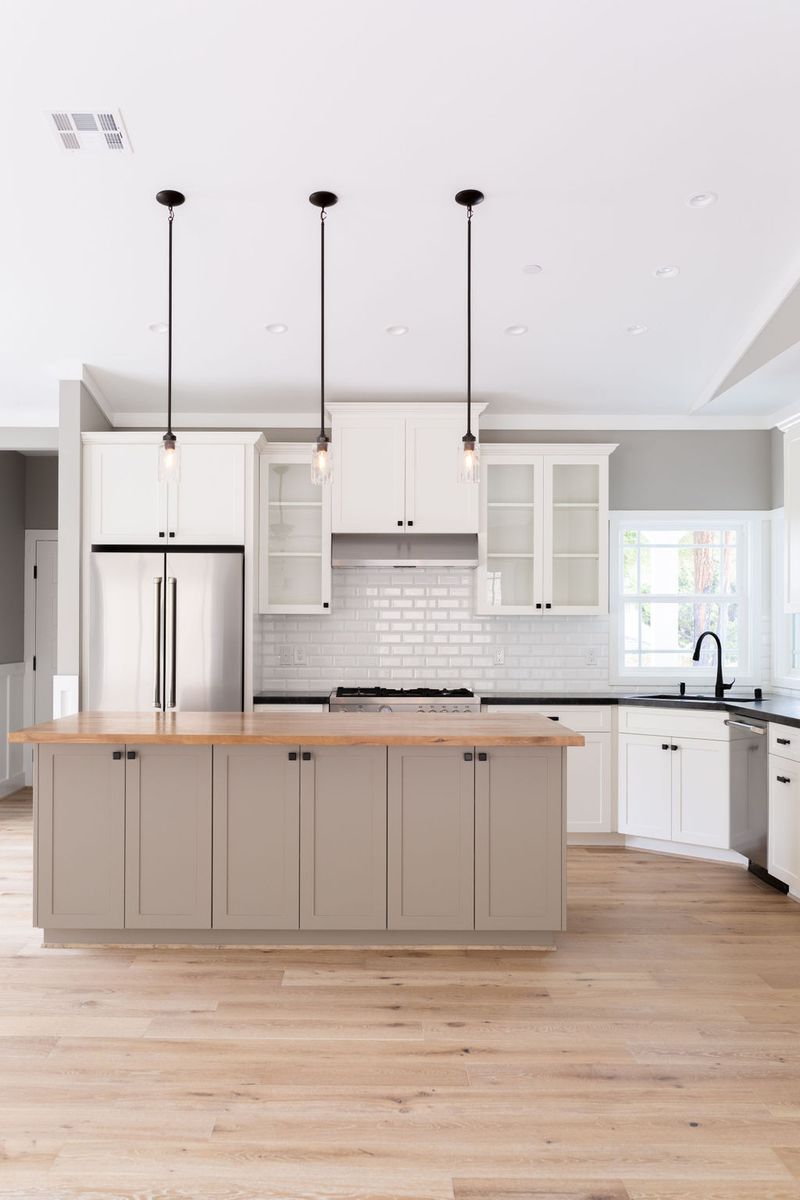 A bright, open kitchen featuring white shaker upper cabinets, a greige center island with butcher block countertop, white subway tile backsplash, and three pendant lights. Light hardwood floors and black hardware complete the transitional design.