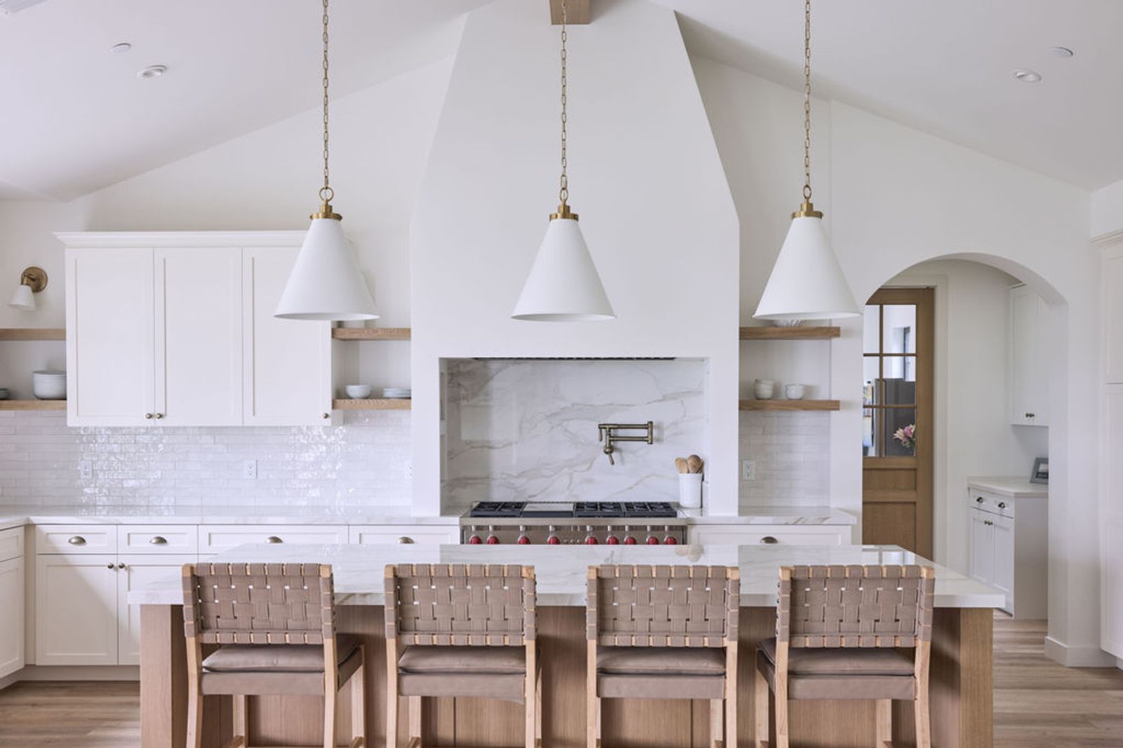 A bright, airy custom kitchen featuring white inset cabinetry, open oak shelving, a dramatic arched plaster range hood with marble slab backsplash, a large island with woven leather bar stools, and three white cone pendant lights with brass hardware.