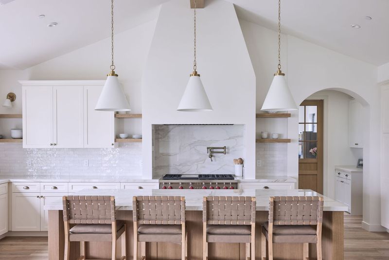 A bright, airy custom kitchen featuring white inset cabinetry, open oak shelving, a dramatic arched plaster range hood with marble slab backsplash, a large island with woven leather bar stools, and three white cone pendant lights with brass hardware.