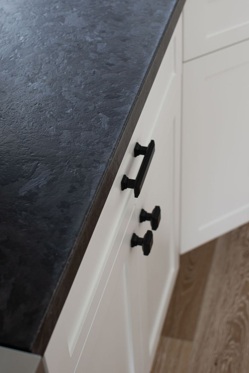 A close-up view of white shaker-style kitchen cabinets with matte black hardware, topped with a dark textured stone countertop and set above light wood flooring.