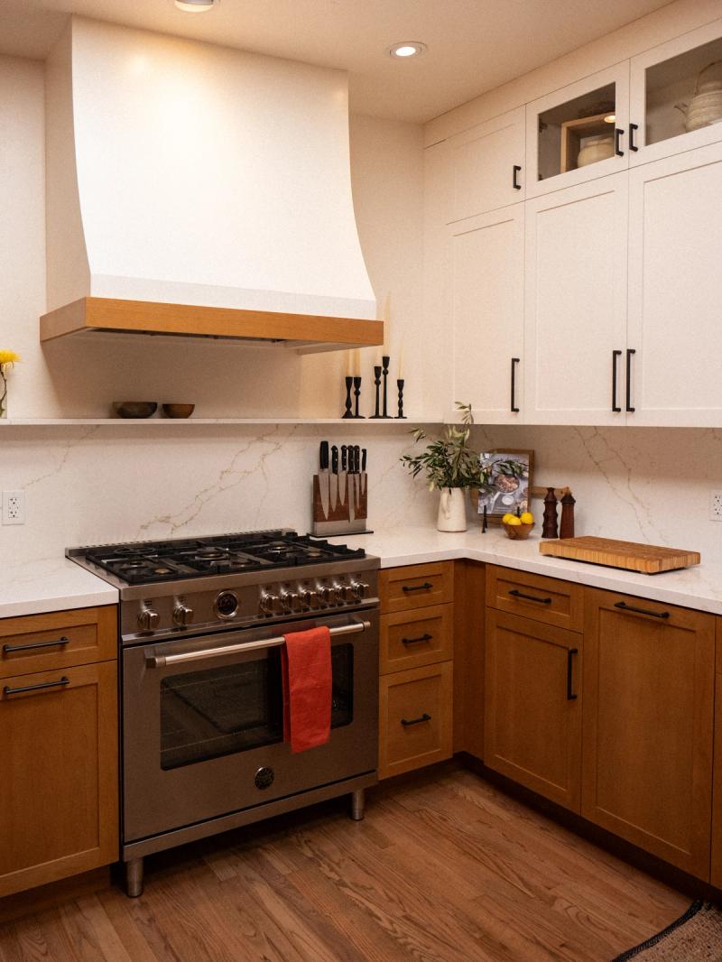 A warm transitional kitchen featuring white upper cabinets, natural wood lower cabinets, a marble-look quartz backsplash, and a professional stainless range beneath a plaster range hood with wood trim.