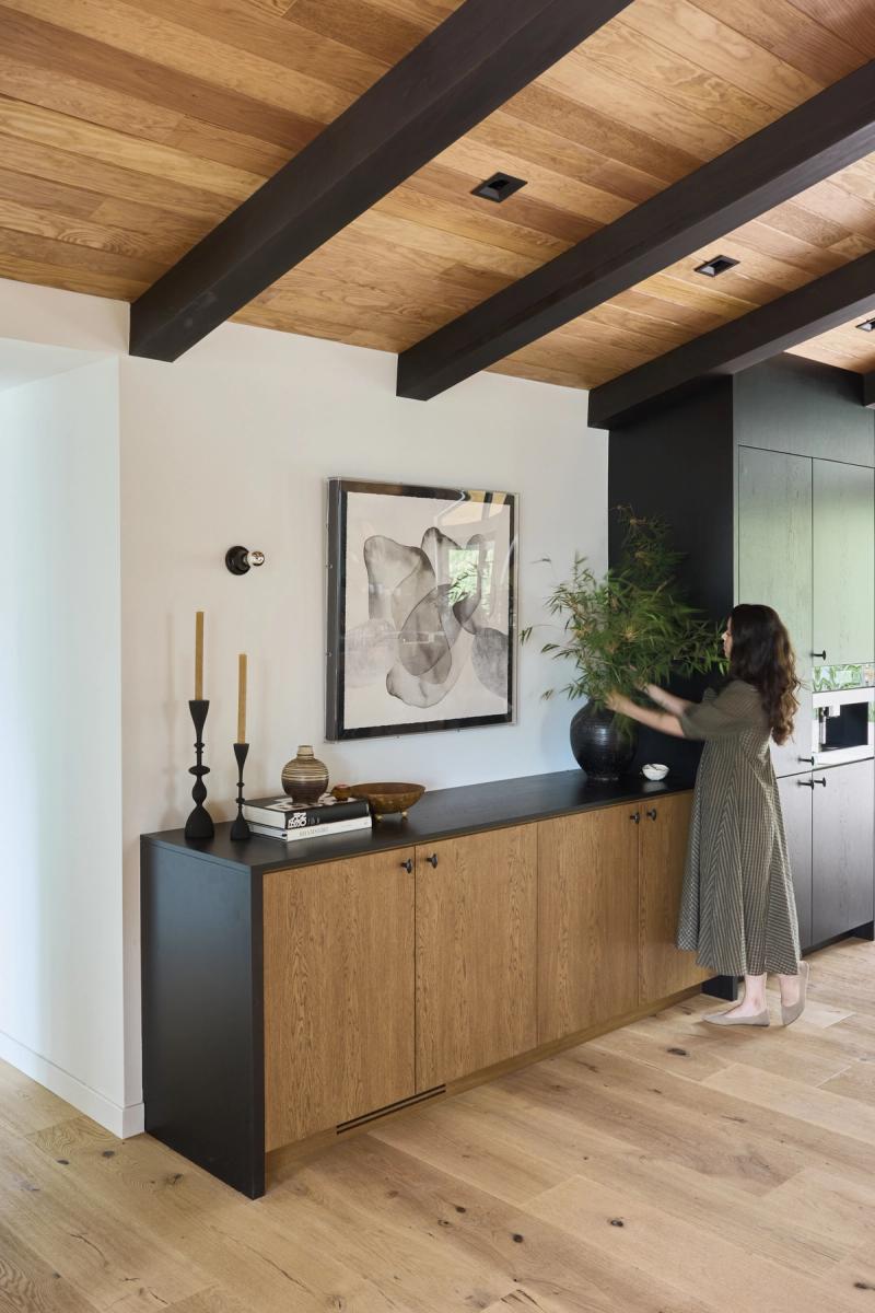 A woman arranges a large plant in a black vase on a floating oak and matte black sideboard cabinet, set against a white wall with abstract artwork, beneath a wood-paneled ceiling with dark beams.