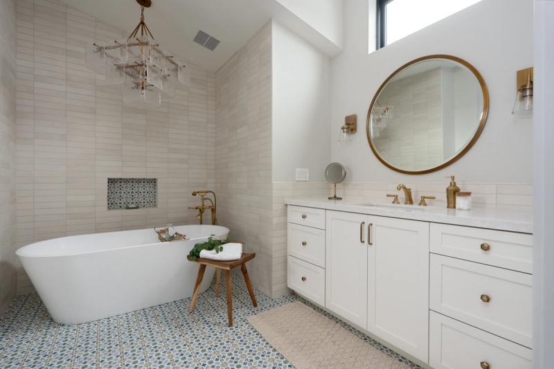 A bright, elegant master bathroom featuring a white shaker-style vanity with brass hardware, a round gold-framed mirror, and a freestanding soaking tub set against white subway tile walls and decorative patterned floor tiles.