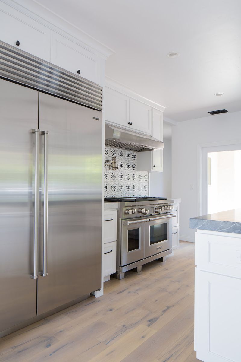 A bright white kitchen featuring a large stainless steel Sub-Zero refrigerator, a Wolf dual-oven range with patterned tile backsplash, white shaker cabinetry, and light hardwood floors.