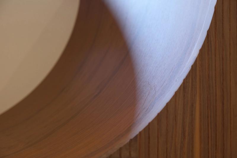 An extreme close-up macro shot of a curved wooden surface with warm brown grain, meeting a white painted or laminate edge, set against a vertical wood panel background.