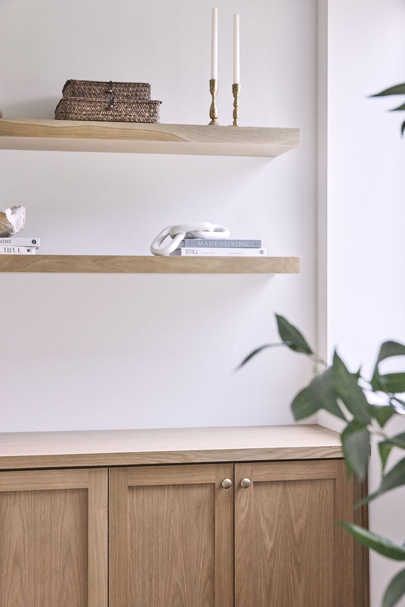 A clean, modern built-in display area featuring light white oak floating shelves styled with books, decorative objects, and brass candleholders, above a matching wood cabinet with inset doors and round knobs.