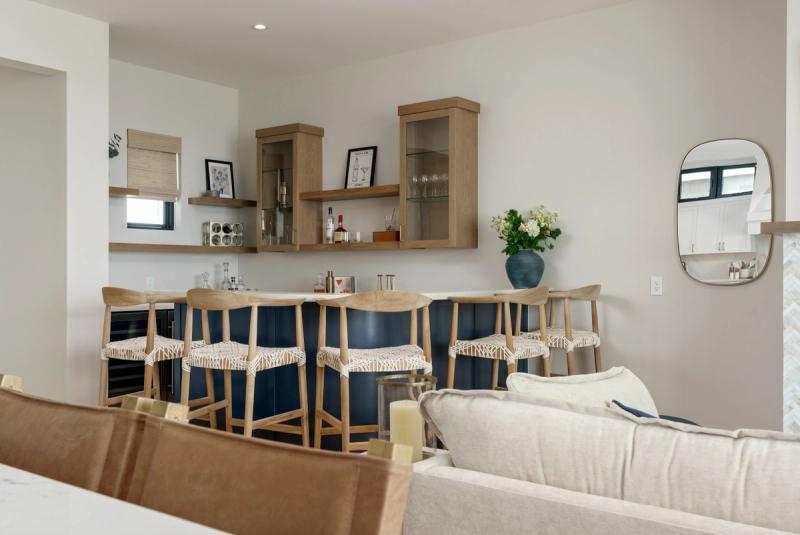 A stylish home bar area featuring navy blue cabinetry, white oak bar stools with woven seats, floating wood shelves, and glass-front upper cabinets displaying barware and spirits.