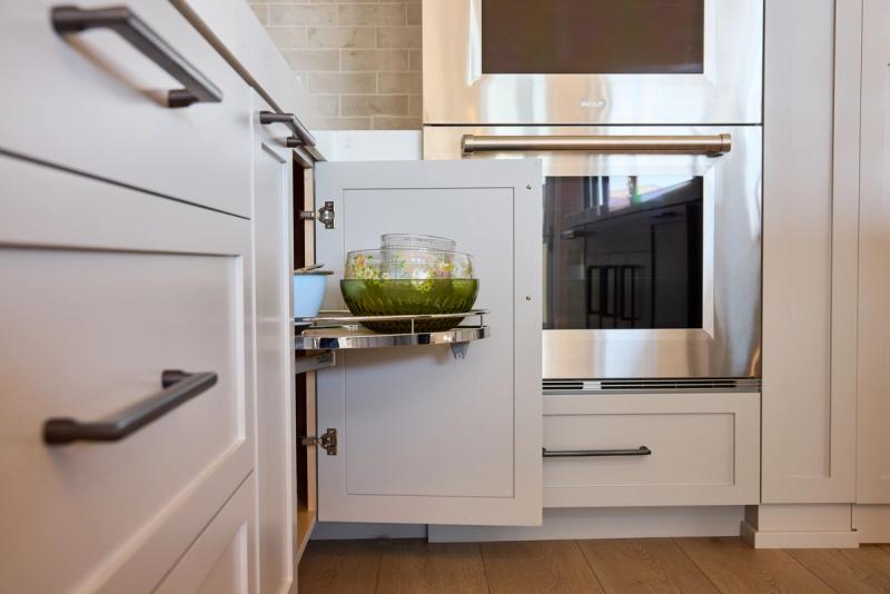A close-up view of an open corner pull-out cabinet in a white shaker-style kitchen, revealing a chrome pull-out shelf holding decorative bowls, positioned next to a stainless steel Wolf double oven.